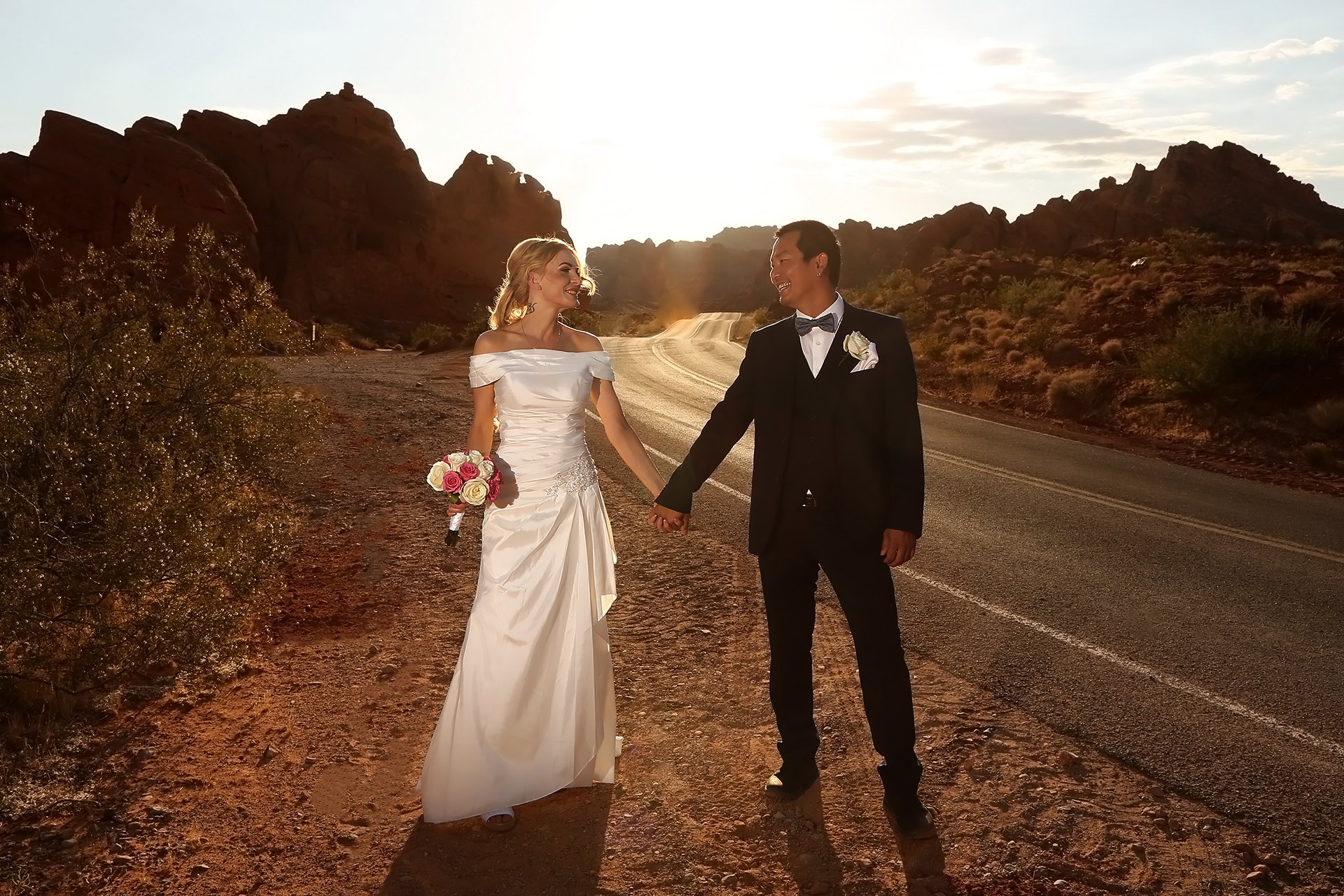 A bride and groom holding hands on a desert road.